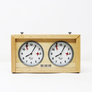 May include: A wooden chess clock with two circular clock faces. Each clock face has a white background, black numerals, and black hands. The clock features the text "GARDE ruhla". The clock is set against a white background.