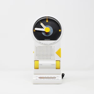 May include: A white Orient AM/FM quartz clock radio with a circular analog clock face featuring black, yellow, and white hands. The device includes a speaker grille, yellow volume and tuning dials, and a horizontal frequency display at the base.