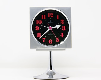 Könnte beinhalten: Ein silberfarbener, quadratischer Wecker mit schwarzem Zifferblatt und roten Ziffern. Die Uhr hat einen Chromfuß und einen weißen pfeilförmigen Stundenzeiger. Auf dem Zifferblatt steht "GARANT" und "2 JEWELS".