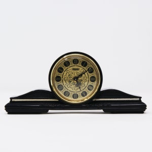 Pu&ograve; includere: Un orologio da mensola nero con un quadrante dorato. Il quadrante presenta numeri romani e decorazioni a volute. L'orologio ha una base curva con un accento argentato.