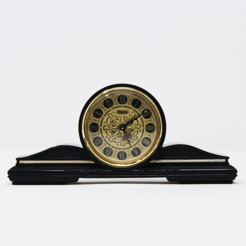 Pu&ograve; includere: Un orologio da mensola nero con un quadrante dorato. Il quadrante presenta numeri romani e decorazioni a volute. L'orologio ha una base curva con un accento argentato.