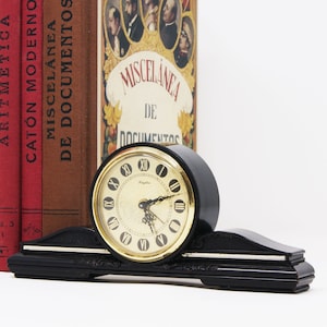 Pu&ograve; includere: Un orologio nero vintage con quadrante dorato e numeri romani. L'orologio &egrave; posto su una base nera con dettagli argentati. Dietro l'orologio ci sono diversi libri antichi con copertine rosse e marroni.