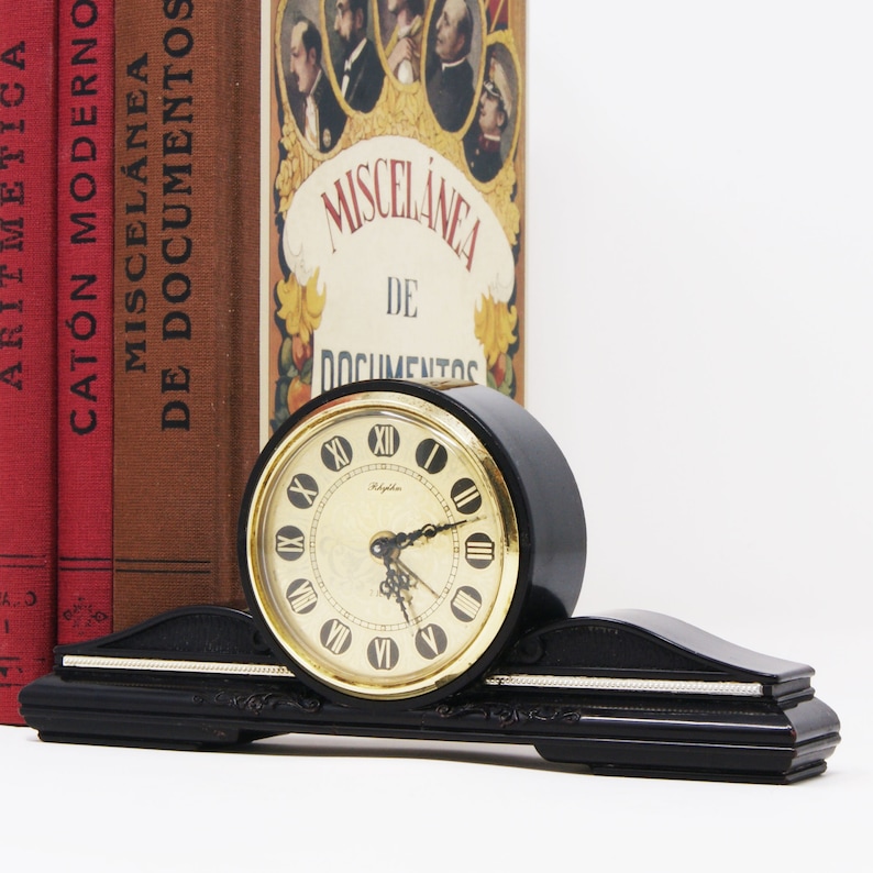 Pu&ograve; includere: Un orologio nero vintage con quadrante dorato e numeri romani. L'orologio &egrave; posto su una base nera con dettagli argentati. Dietro l'orologio ci sono diversi libri antichi con copertine rosse e marroni.