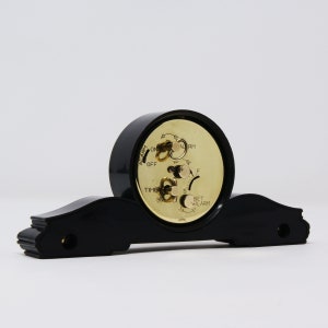 Pu&ograve; includere: Una sveglia vintage nera con quadrante color oro e base decorativa. L'orologio ha manopole per impostare l'ora e la sveglia, con le parole "ALARM", "TIME" e "SET ALARM" visibili. L'orologio &egrave; posto su uno sfondo bianco.