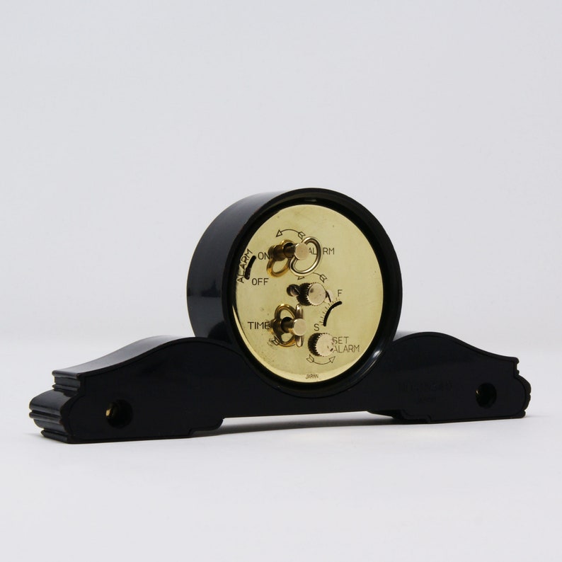 Pu&ograve; includere: Una sveglia vintage nera con quadrante color oro e base decorativa. L'orologio ha manopole per impostare l'ora e la sveglia, con le parole "ALARM", "TIME" e "SET ALARM" visibili. L'orologio &egrave; posto su uno sfondo bianco.