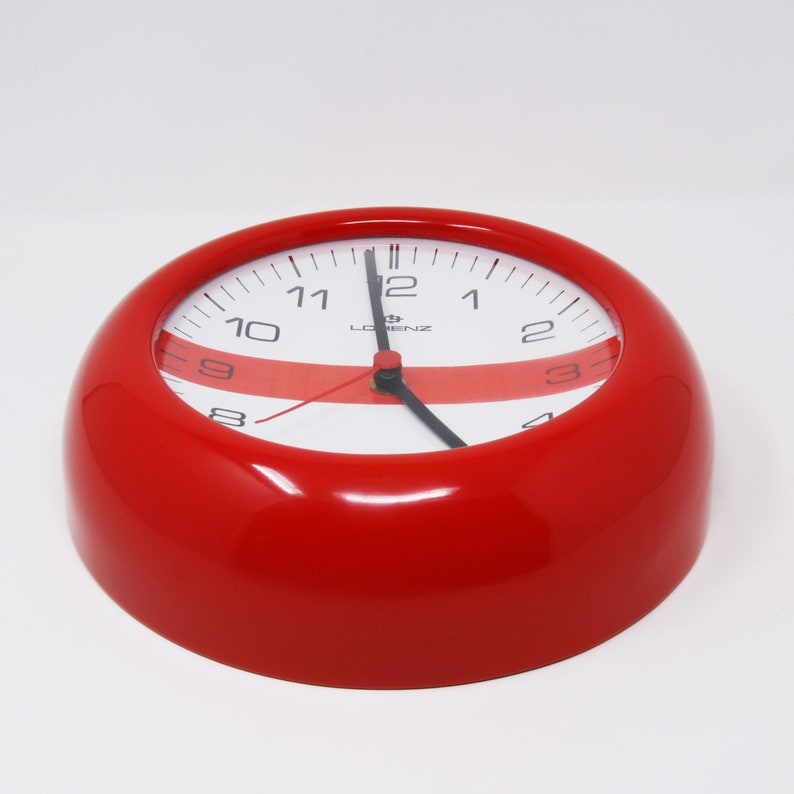 May include: A red, round clock with white face and black numbers. The clock has a red stripe across the face and the brand name "LORENZ" is printed on the face.