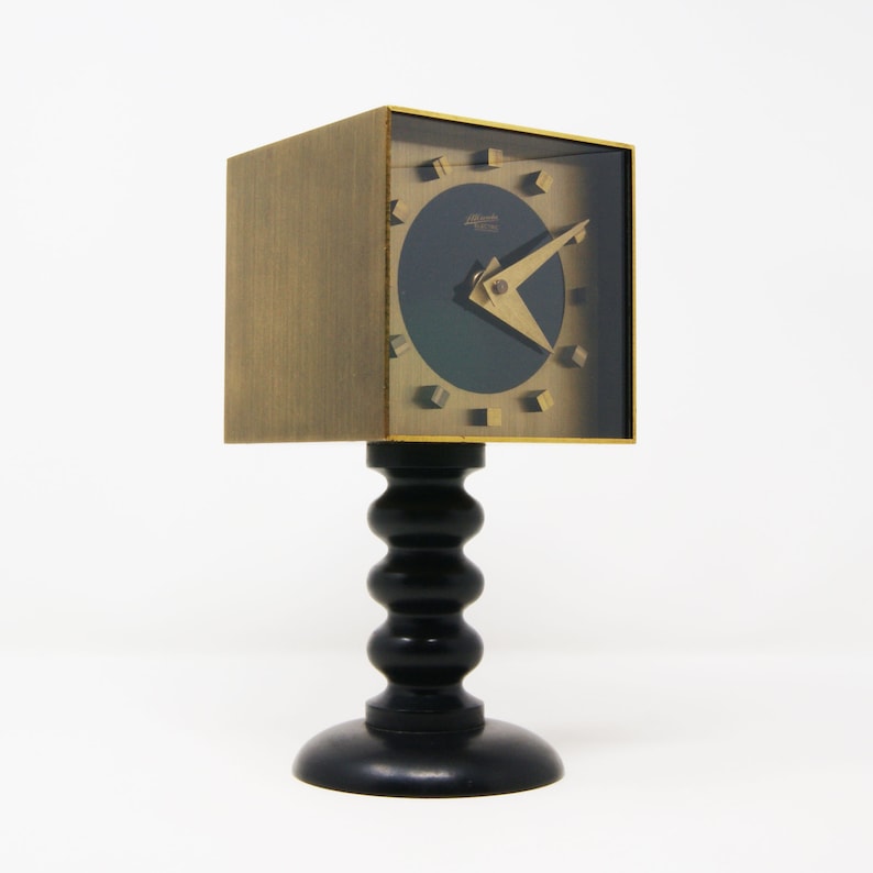 May include: A gold-toned clock with a black face and a unique design. The clock is on a black stand with a rounded base.