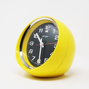 Peut inclure: Un réveil jaune avec un cadran noir et des accents argentés. Le cadran de l'horloge a des chiffres noirs et une aiguille des secondes rouge. L'horloge indique "Jerger" et "Made in Germany".