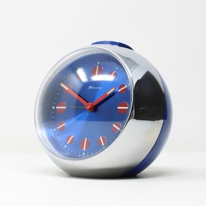 May include: A blue and silver alarm clock with red hands and white accents. The clock face has the word "Blessing" printed on it.