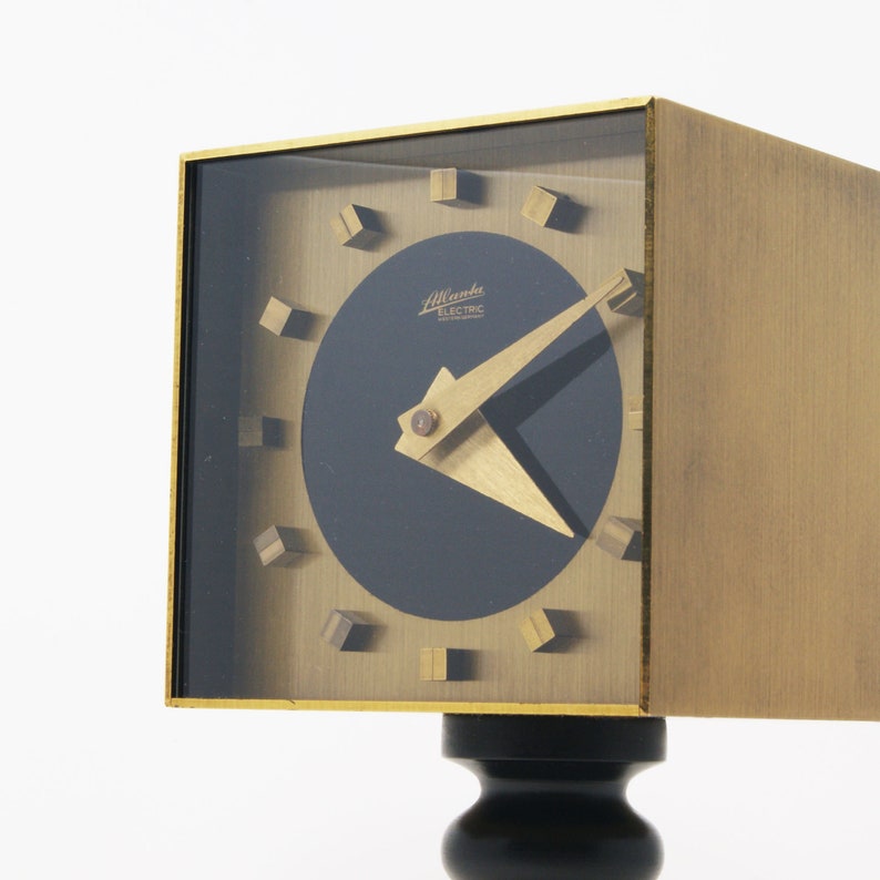 May include: A gold-toned square clock with a black face and gold hands. The clock has a unique design with small squares around the edge of the face. The clock is mounted on a black pedestal.