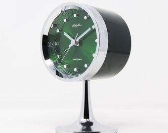 Könnte beinhalten: Ein Chrom-Wecker mit grünem Zifferblatt. Die Uhr hat ein weißes Zifferblatt mit schwarzen Zahlen und Zeigern. Die Uhr steht auf einem Chromständer.