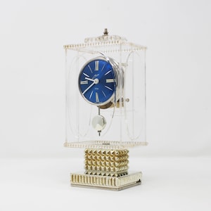 Könnte beinhalten: Eine vintage, transparente Acryluhr mit blauem Zifferblatt und silbernen Zeigern. Die Uhr hat ein Pendel und ist auf einem goldfarbenen Sockel montiert.