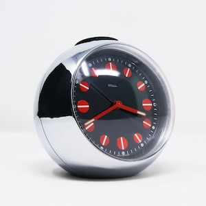May include: A silver alarm clock with a black face and red numbers. The clock has a round, chrome-plated case and a clear glass face. The clock is made in West Germany.
