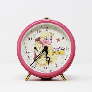 Puede incluir: Reloj despertador rosa con una esfera blanca que presenta una ilustración de Barbie y el texto "Barbie". El reloj tiene agujas doradas y una perilla dorada.