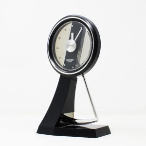 Puede incluir: Un reloj de escritorio Rhythm Quartz negro y plateado. El reloj tiene una esfera redonda con un centro plateado y detalles en negro. El reloj está sostenido por una base negra y un soporte plateado. La esfera del reloj tiene el texto "Rhythm Quartz".