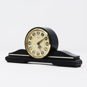 Pu&ograve; includere: Un orologio da mensola nero con quadrante color oro e numeri romani. L'orologio ha una base curva con finiture argentate e la parola "Rhythm" &egrave; stampata sul quadrante.