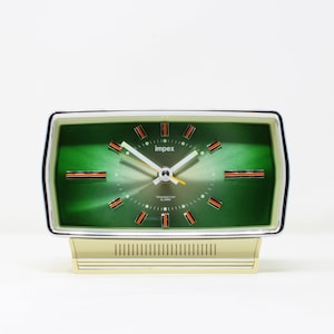 May include: A green and gold vintage alarm clock with a white face and black hands. The clock has a silver trim and the text "Impex Transistor Alarm" on the face.