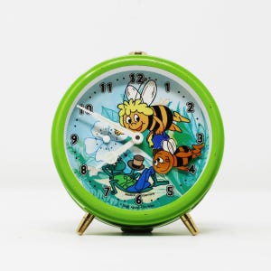 Könnte beinhalten: Ein grüner, vintage Wecker mit einem Cartoon-Bienen- und Frosch-Design. Das Zifferblatt hat einen hellblauen Hintergrund mit weißen Zeigern und goldfarbenen Beinen. Die Uhr ist mit "Made in Germany" gekennzeichnet.