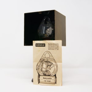 May include: A close-up of a clock movement with a brown wooden box. The clock movement is labeled "KIENZLE" and "Babyzelle 1.5 Volt".