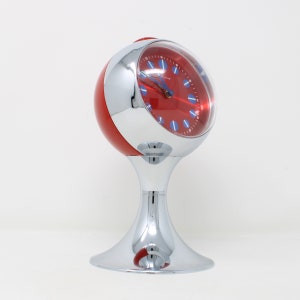 Puede incluir: Un reloj despertador rojo y cromado con una esfera redonda y un soporte. La esfera del reloj tiene números y manecillas azules.