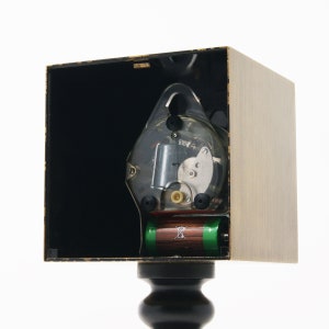 May include: A clock mechanism with a clear plastic cover, showing the inner workings of the clock. The clock is mounted in a wooden box with a black interior. Two batteries are visible at the bottom of the box.