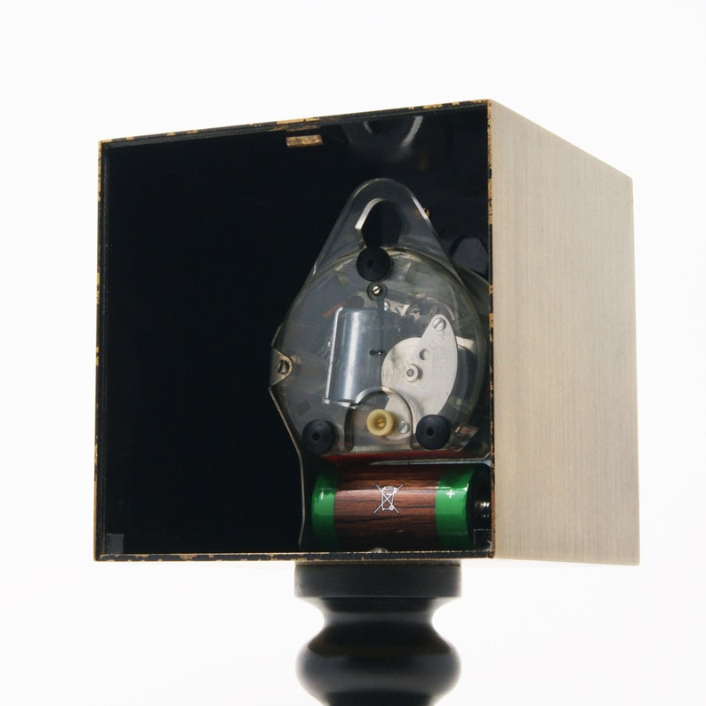 May include: A clock mechanism with a clear plastic cover, showing the inner workings of the clock. The clock is mounted in a wooden box with a black interior. Two batteries are visible at the bottom of the box.
