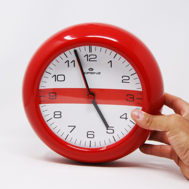 May include: A red round wall clock with white face and black hands. The clock has a red and white stripe design and the brand name "ORENZ" is printed on the face.