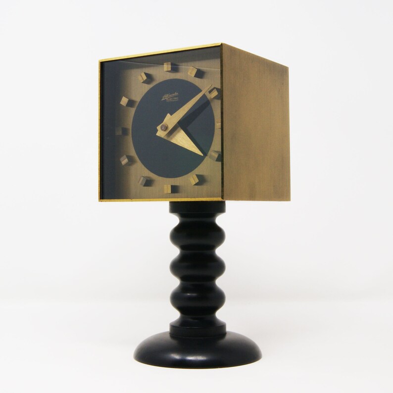 May include: A gold-toned clock with a black face and a unique design. The clock is mounted on a black, fluted pedestal.
