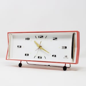 Könnte beinhalten: Vintage rechteckiger Wecker mit rotem Rahmen und weißem Zifferblatt. Die Uhr hat schwarze Ziffern, goldene Zeiger und den Markennamen "Rhythm". Sie wird von einem silbernen Metallständer mit schwarzen Füßen getragen.