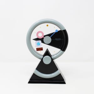 Puede incluir: Un reloj Rhythm Japan de estilo retro con una base triangular negra y gris y una esfera circular. La esfera presenta un fondo blanco con una sección negra y gris, un círculo rosa y detalles en azul y gris.