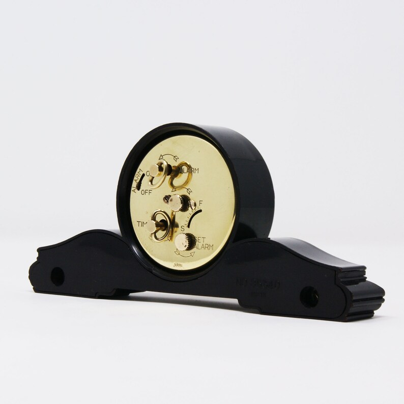 Pu&ograve; includere: Una sveglia vintage nera con quadrante e manopole dorate. L'orologio ha una base curva e un quadrante rotondo. Sul quadrante sono stampate le parole "ALARM", "TIME" e "SET ALARM".