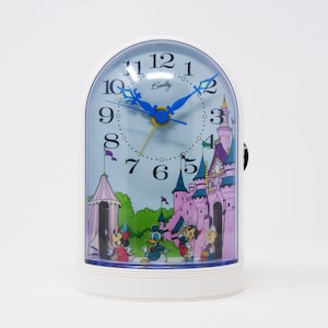 以下が含まれることがあります： 白地に青とピンクのディズニー城のシーンが描かれた目覚まし時計。時計の文字盤は青で、白い数字とブラッドリーロゴが書かれている。ミッキーマウス、ドナルドダック、その他のディズニーキャラクターがシーンに登場している。