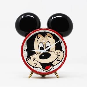 May include: Red Mickey Mouse alarm clock with black ears and a white face. The clock face features Mickey Mouse's face with the words "Mickey Mouse" and numbers 1 through 12.