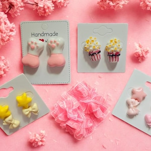 May include: A collection of handmade earrings on white cards, including paw prints, cupcakes, stars, and bows. The earrings are in shades of pink, yellow, and white, arranged on a pink surface with floral accents. The word "Handmade" is visible on one card.