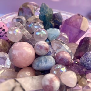 May include: A collection of assorted polished gemstones and crystals in a clear glass bowl. The assortment includes spherical, heart-shaped, and raw-cut stones in shades of pink, purple, blue, and clear, with a small pink tag reading Spell Bin.