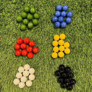 Peut inclure: Assortiment de billes de verre colorées en vert, bleu, rouge, jaune, blanc et noir. Les billes sont disposées en six groupes sur une surface herbeuse verte.