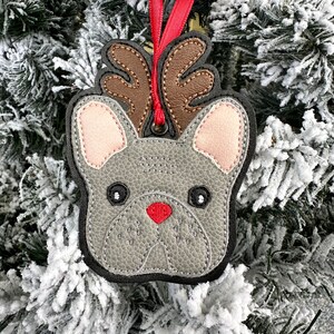 May include: A gray felt French bulldog ornament with brown antlers and a red nose, hanging from a red ribbon on a snowy Christmas tree.