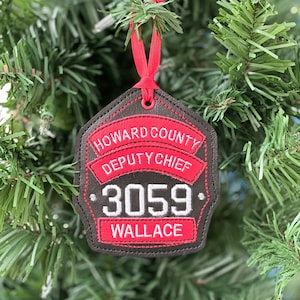 May include: A decorative Christmas ornament shaped like a badge, featuring the text "HOWARD COUNTY DEPUTY CHIEF" and "3059 WALLACE" in white and red on a black background. The ornament is hanging from a red ribbon and is set against a backdrop of green Christmas tree branches.
