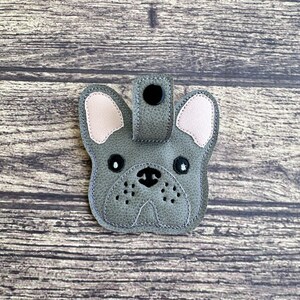以下が含まれることがあります： ピンクの耳と黒い目をしたグレーのフェルト製のフレンチブルドッグのキーホルダー。キーホルダーの上部に黒いボタンが付いています。