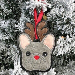 May include: A gray felt French bulldog ornament with brown antlers and a red nose, hanging from a red ribbon on a snowy Christmas tree.