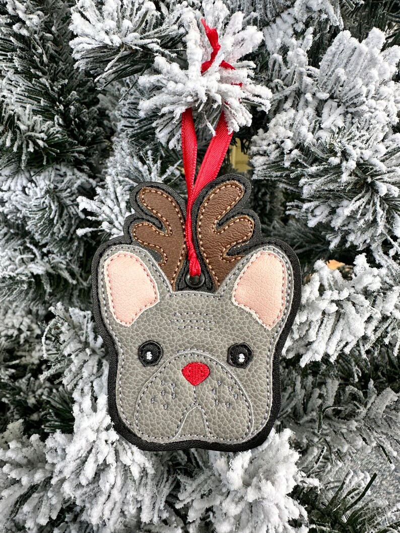 May include: A gray felt French bulldog ornament with brown antlers and a red nose, hanging from a red ribbon on a snowy Christmas tree.