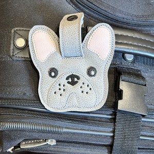 以下が含まれることがあります： ピンクの耳をしたフレンチブルドッグの形をしたグレーの革製の荷物タグ。タグは黒い目と黒い鼻をしています。