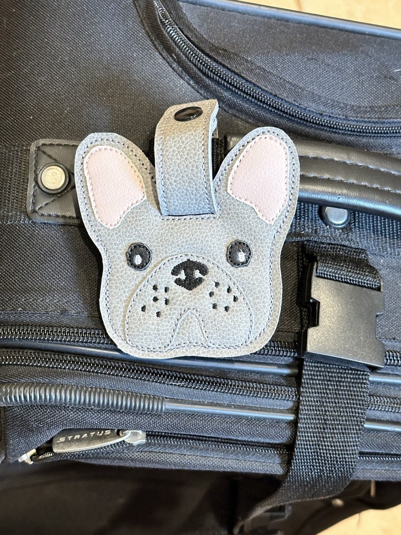 以下が含まれることがあります： ピンクの耳をしたフレンチブルドッグの形をしたグレーの革製の荷物タグ。タグは黒い目と黒い鼻をしています。