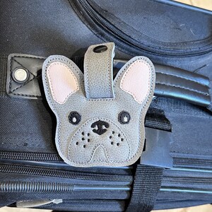 以下が含まれることがあります： 黒の旅行鞄に付けられた、グレーとピンクのフレンチブルドッグの荷物タグ。黒い目と黒い鼻が特徴です。