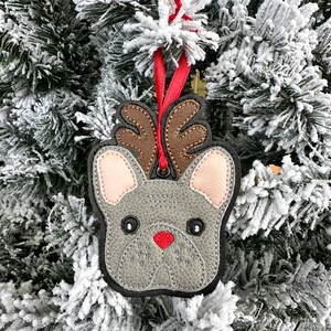 May include: A gray felt French bulldog ornament with brown antlers and a red nose. The ornament is hanging on a snowy Christmas tree.