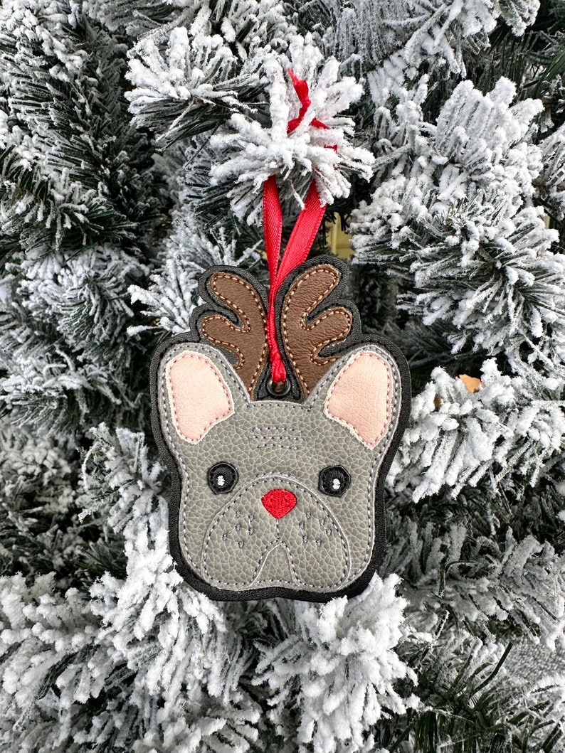 May include: A gray felt French bulldog ornament with brown antlers and a red nose. The ornament is hanging on a snowy Christmas tree.