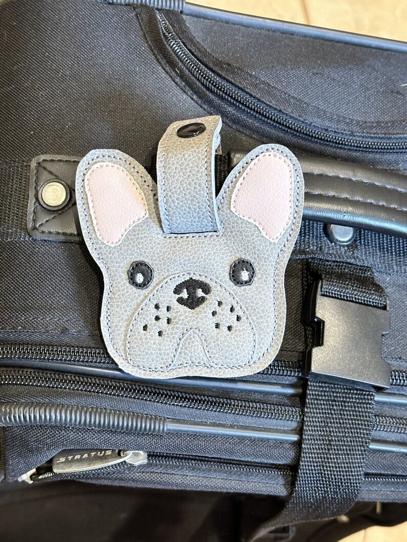 以下が含まれることがあります： 黒の旅行鞄に付けられた、グレーとピンクのフレンチブルドッグの荷物タグ。黒い目と黒い鼻が特徴です。