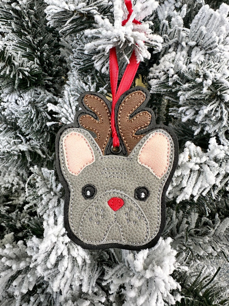 May include: A gray felt French bulldog ornament with brown antlers and a red nose, hanging from a red ribbon on a snowy Christmas tree.