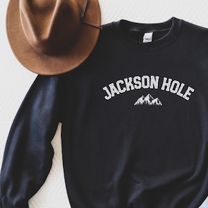 Peut inclure: Sweat-shirt noir avec un texte blanc qui dit "JACKSON HOLE" et un graphique de montagnes.