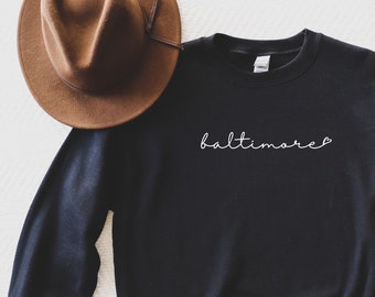 May include: Black sweatshirt with the word "baltimore" written in white cursive on the front. The sweatshirt is laid flat on a white surface with a brown hat and blue jeans.